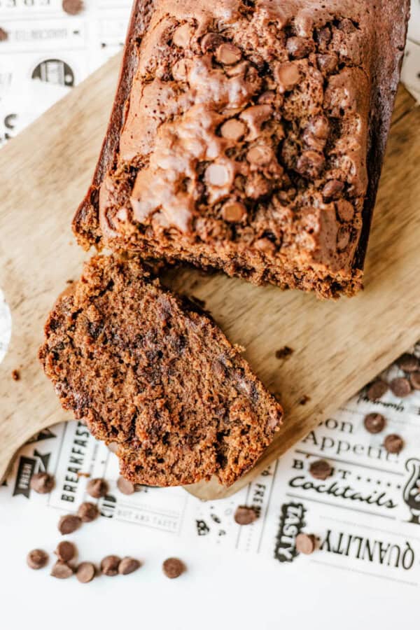 A loaf of chocolate chip banana bread sits on a wooden board with one slice partially cut, reminiscent of classic chocolate zucchini bread. Chocolate chips are visible inside and scattered around, against a backdrop of patterned food-themed paper.