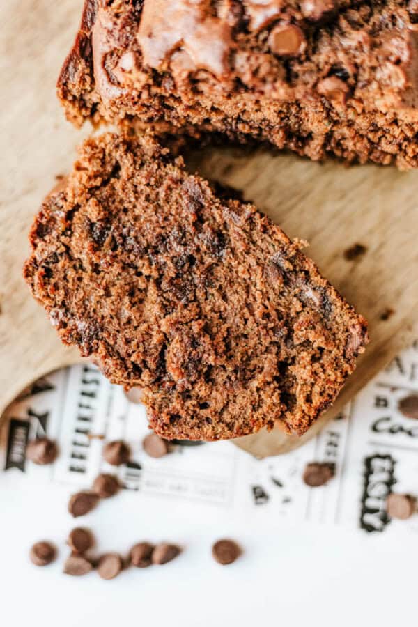 A close-up of a sliced loaf of chocolate chip banana bread and chocolate zucchini bread on a wooden board, with a few chocolate chips scattered nearby on printed paper.