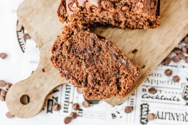 A sliced loaf of chocolate zucchini bread sits on a wooden cutting board, with one slice in front displaying its moist texture and chocolate bits. Extra chocolate chips are scattered on a printed paper underneath.