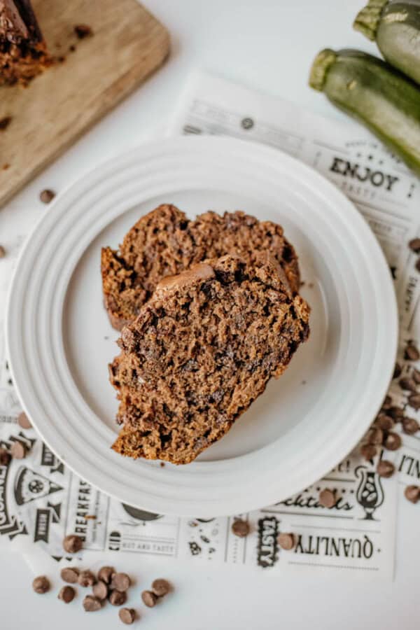 Two slices of chocolate zucchini bread rest on a white plate, with chocolate chips scattered around and fresh zucchini in the background atop a patterned newspaper-style surface.