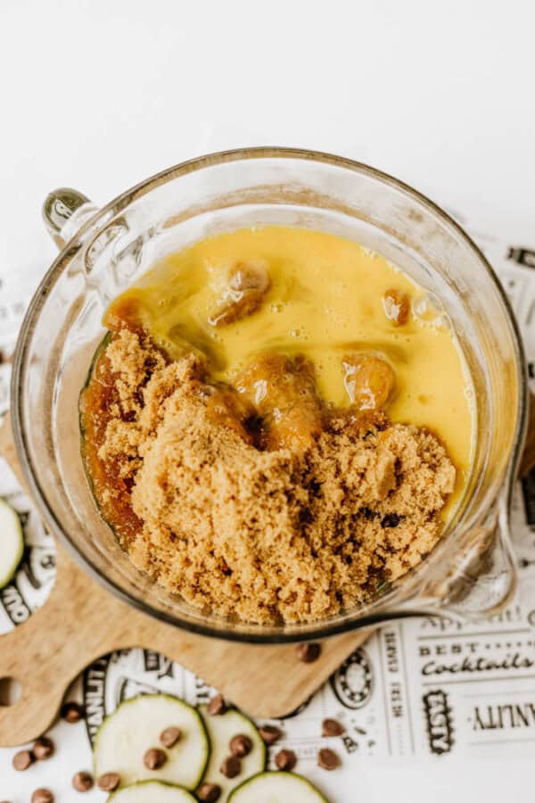 A glass mixing bowl filled with whisked eggs, brown sugar, and other baking ingredients sits on a counter next to chocolate chips and zucchini slices—perfect for making chocolate zucchini bread.