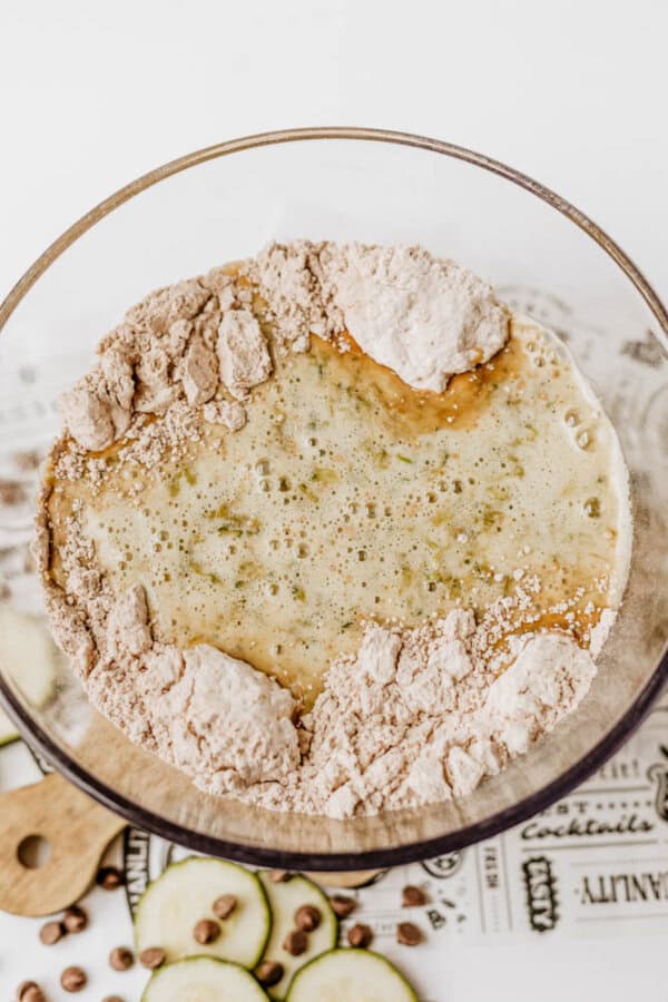 A glass mixing bowl containing flour and wet ingredients, partially combined for chocolate zucchini bread, sits on a wooden board with sliced zucchini and chocolate chips nearby.