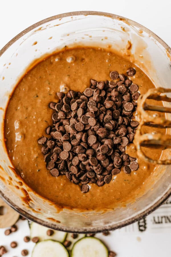 A mixing bowl filled with chocolate zucchini bread batter and topped with a pile of chocolate chips. An electric mixer beater is partially visible on the right side of the bowl.