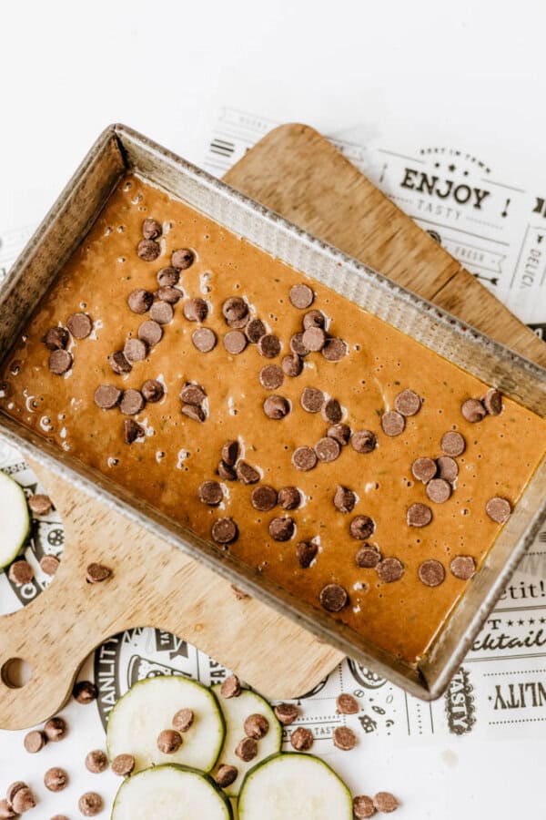 A metal loaf pan filled with chocolate zucchini bread batter sits on a wooden cutting board, topped with chocolate chips and surrounded by fresh zucchini slices.
