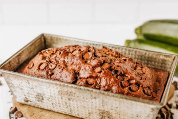 A loaf of chocolate zucchini bread in a metal baking pan, its golden brown top studded with chocolate chips. Blurred zucchinis are visible in the background.
