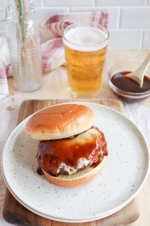 A cooked hamburger covered in melted white cheese and BBQ sauce on a white bun on a cream speckled plate with a glass of beer in the background.