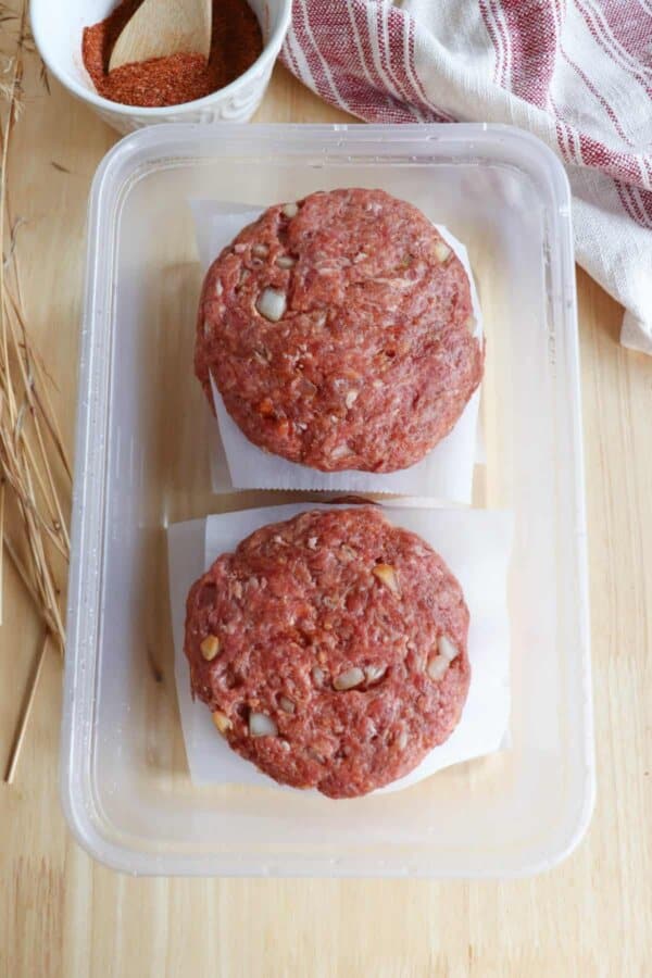 Raw hamburger patties stacked between sheets of parchment paper ready to be frozen in a clear plastic container.