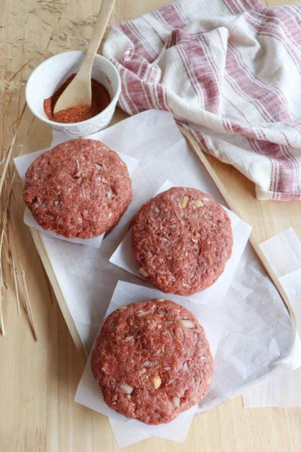 Raw hamburger patties stacked between sheets of parchment paper on a wooden table with a red and white striped dish cloth.