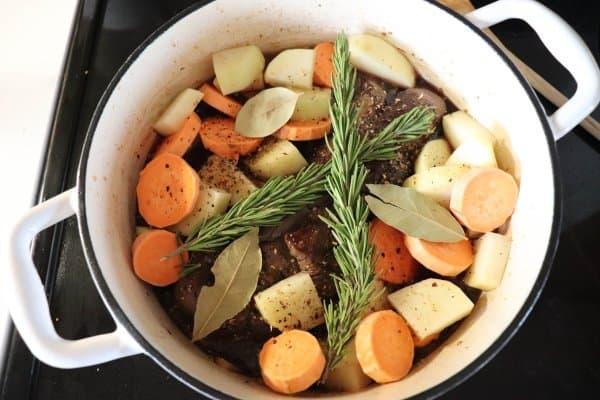 Venison roast in a white dutch oven with raw carrots, potatoes, rosemary and bay leaves.