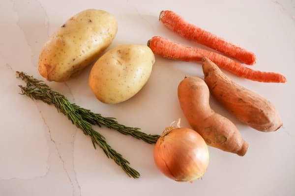 Potatoes, carrots, onion, yams and rosemary on a white counter.