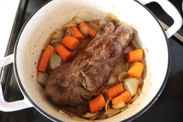 Venison roast in a white dutch oven with carrots and onion.
