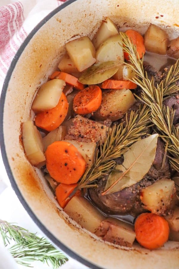 Venison roast in a white dutch oven with carrot, potatoes, rosemary and bay leaves.