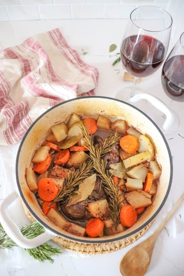 Venison roast in a white dutch oven with carrots, potatoes, rosemary and bay leaves.