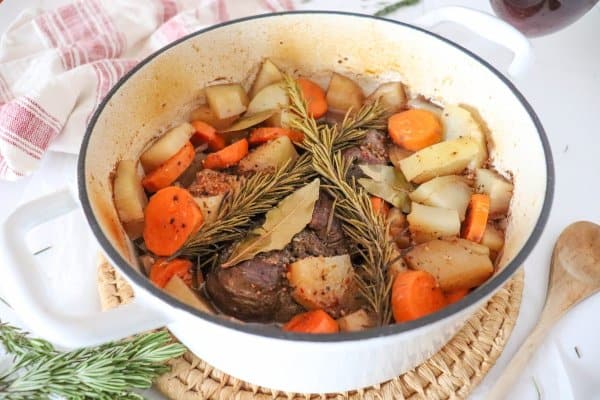 Venison roast in a white dutch oven with carrots, potatoes, rosemary and bay leaves.