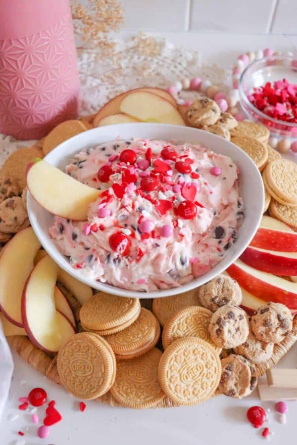 An apple is being dipped into the Valentine's Day dessert dip recipe that is pink with chocolate chips and topped with red M&Ms and heart sprinkles.