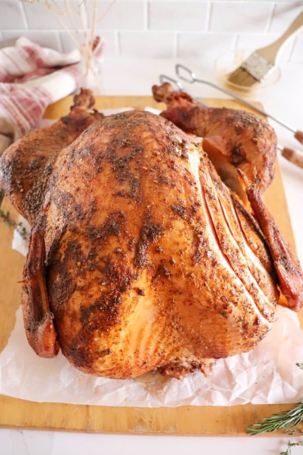 Golden-brown roasted turkey on a wooden cutting board, resting on parchment paper—crafted to perfection with a Traeger Smoked Turkey Recipe. Herbs and a baking brush linger in the background, enhancing the culinary scene.