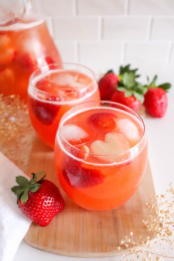 Two glasses of pink strawberry drink on a wooden board, garnished with fresh strawberries and ice cubes. A pitcher filled with the same Valentine's Day punch recipe is in the background, surrounded by whole strawberries and delicate flowers.