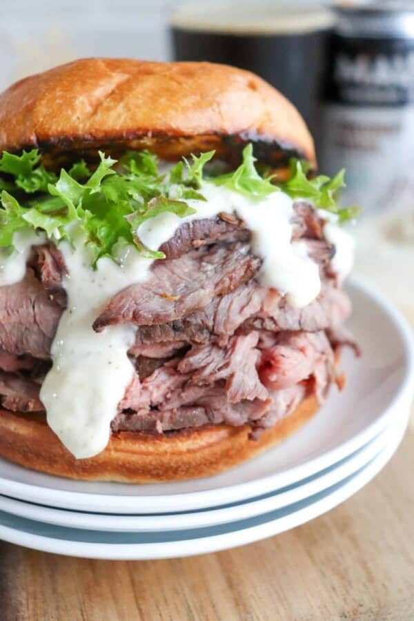 Close-up of a delectable roast beef sandwich, crafted from a classic recipe, with leafy greens and creamy sauce, served on a fresh bun. The sandwich rests on a stack of white plates with a blurred beverage can in the background.