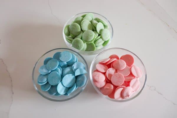 Blue, green and pink chocolate candy melts in three glass bowls.