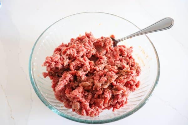 Ground hamburger meat mixed with spices in a large clear glass bowl.