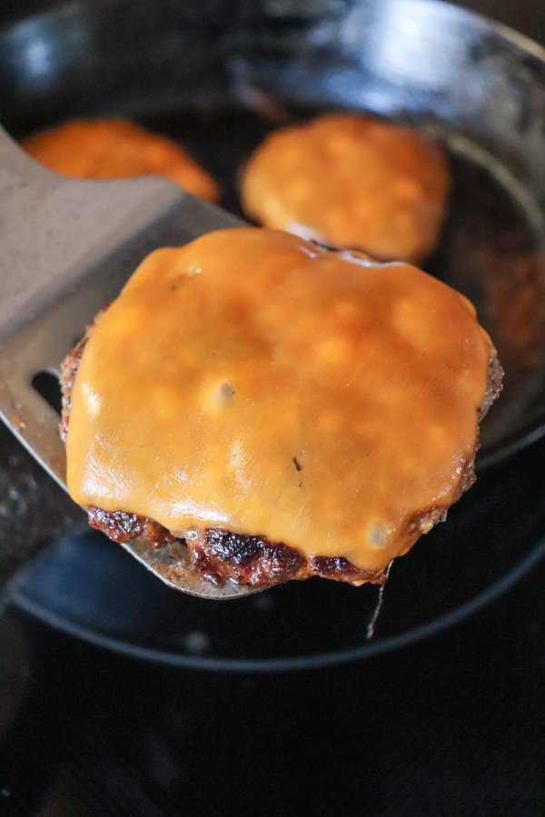 Cooked burger topped with melted orange cheese on a metal spatula with a cast iron skillet in the background.
