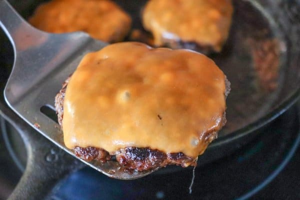 Cooked burger topped with melted orange cheese on a metal spatula with a cast iron skillet in the background.