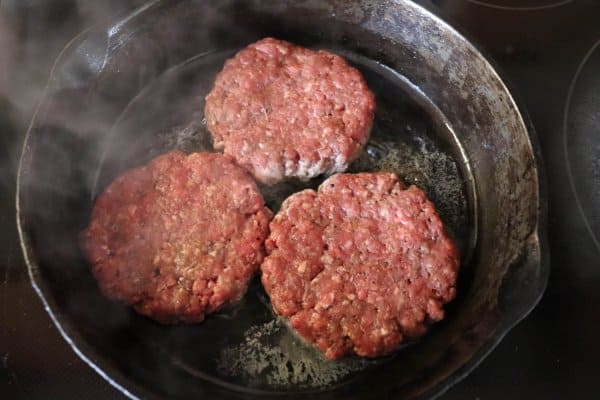 Three raw hamburger patties in a cast  iron skillet with steam rising.