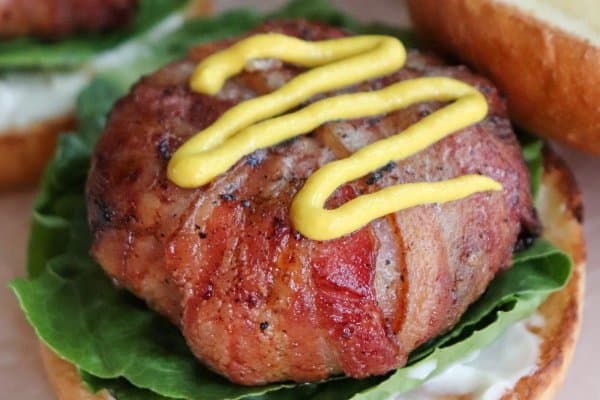 Burger wrapped in bacon with a squiggle of yellow mustard on top on a bed of lettuce with mayo on a toasted white hamburger bun.