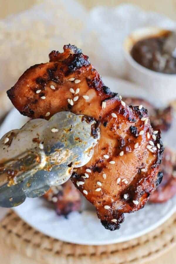 Tongs are holding a grilled teriyaki Chicken thigh.
