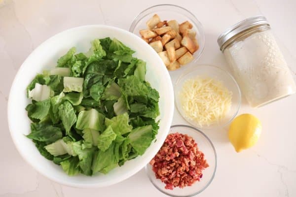 Chopped romaine lettuce, bacon bits, shredded parmesan cheese, lemon croutons, and a mason jar of salad dressing.