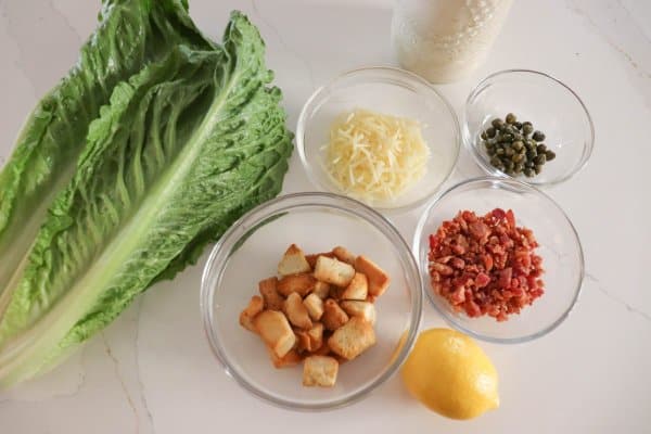 Classic Caesar salad recipe  ingredients laid out on a white surface, romaine lettuce, shredded parmesan cheese, capers, croutons, bacon bits, and a lemon.