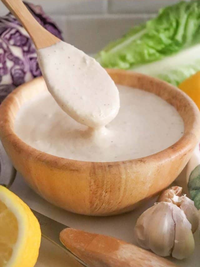 A wooden bowl filled with creamy white dressing, with a wooden spoon above it. Surrounding the bowl are fresh lemon, garlic, and lettuce. Purple cabbage is visible in the background, suggesting a fresh and healthy setting.