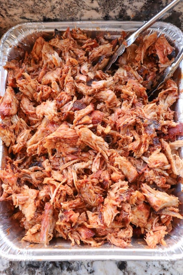 Smoked Pulled Pork close up in an aluminum tray covered in bbq sauce with metal tongs.