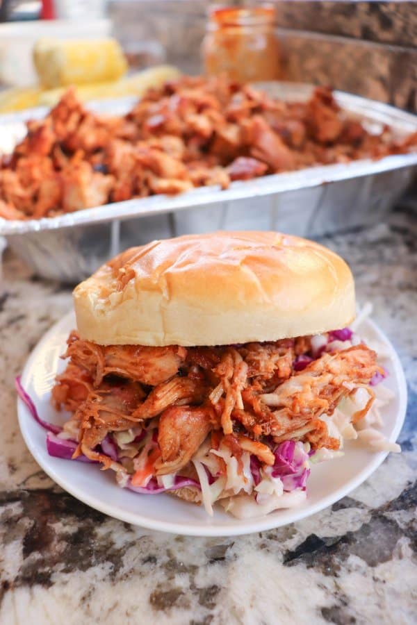 Smoked pulled pork on a white bun with coleslaw on a white plate with a tray of pulled pork in the background.