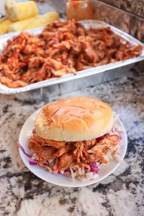 Smoked pulled pork on a white bun with coleslaw on a white plate with a tray of pulled pork in the background.