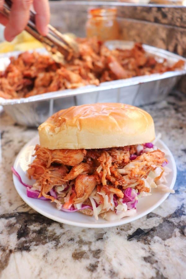 Smoked pulled pork on a white bun with coleslaw on a white plate with a tray of pulled pork in the background.