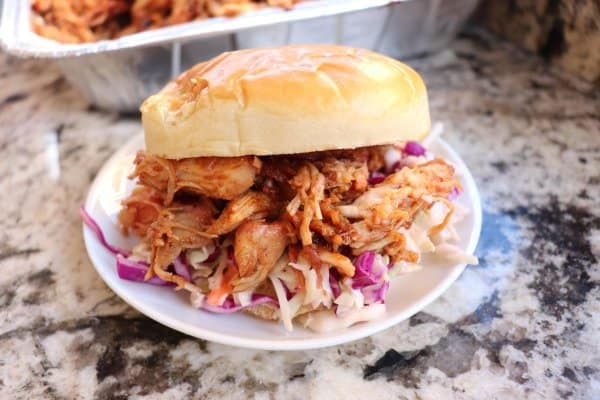 Smoked pulled pork on a white bun with coleslaw on a white plate with a tray of pulled pork in the background.