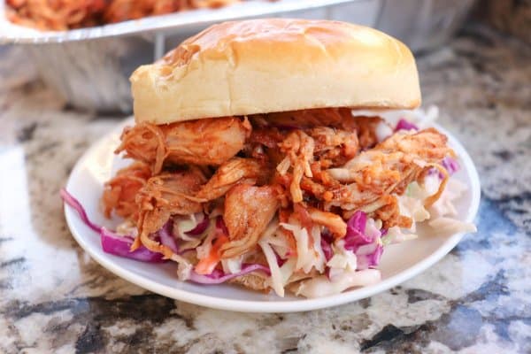Smoked pulled pork on a white bun with coleslaw on a white plate with a tray of pulled pork in the background.