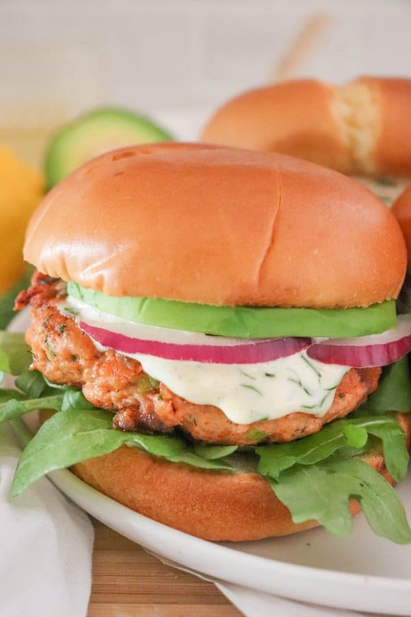 Salmon burger topped with creamy dill sauce, red onion and avocado on a bed of lettuce in a white bun.