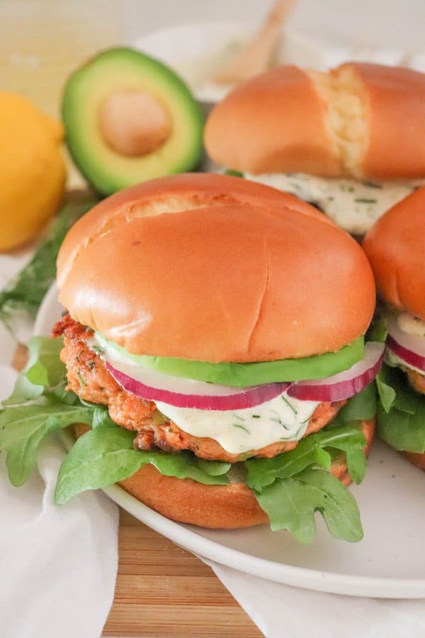 Salmon burger topped with creamy dill sauce, red onion and avocado on a bed of lettuce in a white bun.