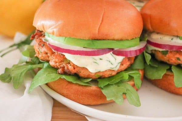 Salmon burger topped with creamy dill sauce, red onion and avocado on a bed of lettuce in a white bun.
