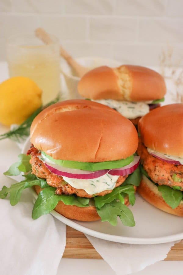 Salmon burger topped with creamy dill sauce, red onion and avocado on a bed of lettuce in a white bun.