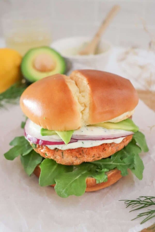 Salmon burger topped with creamy dill sauce, red onion and avocado on a bed of lettuce in a white bun.