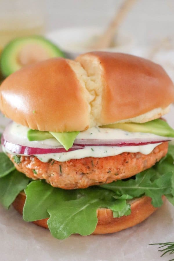 Salmon burger topped with creamy dill sauce, red onion and avocado on a bed of lettuce in a white bun.