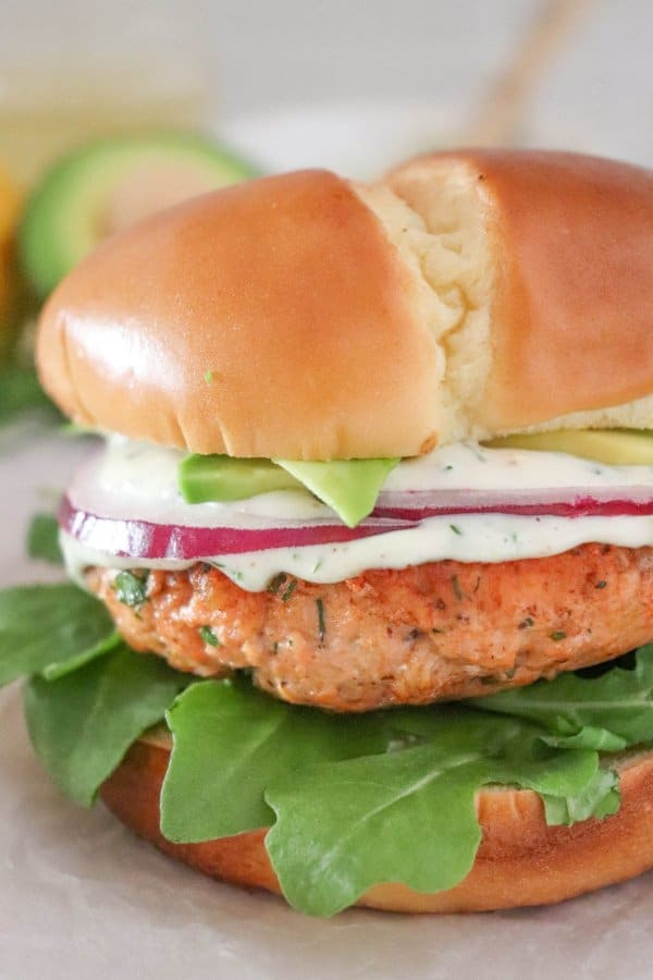 Close up of salmon burger topped with creamy dill sauce, red onion and avocado on a bed of lettuce in a white bun.
