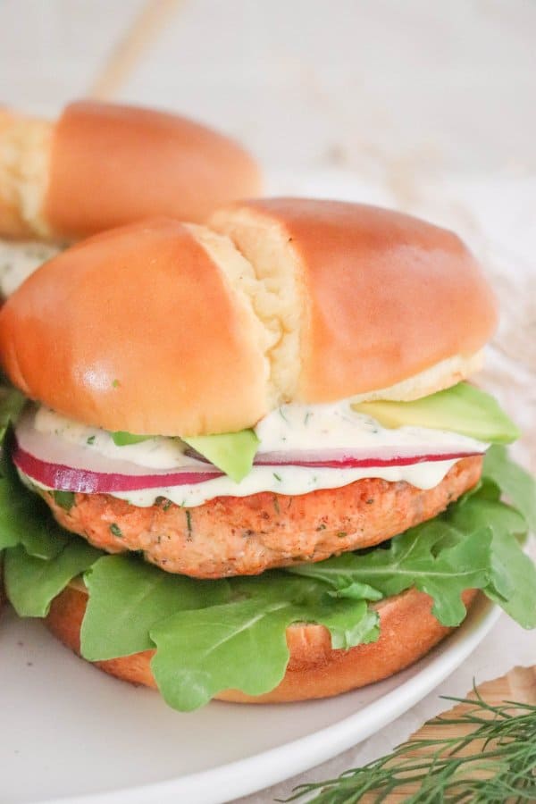 Salmon burger topped with creamy dill sauce, red onion and avocado on a bed of lettuce in a white bun.