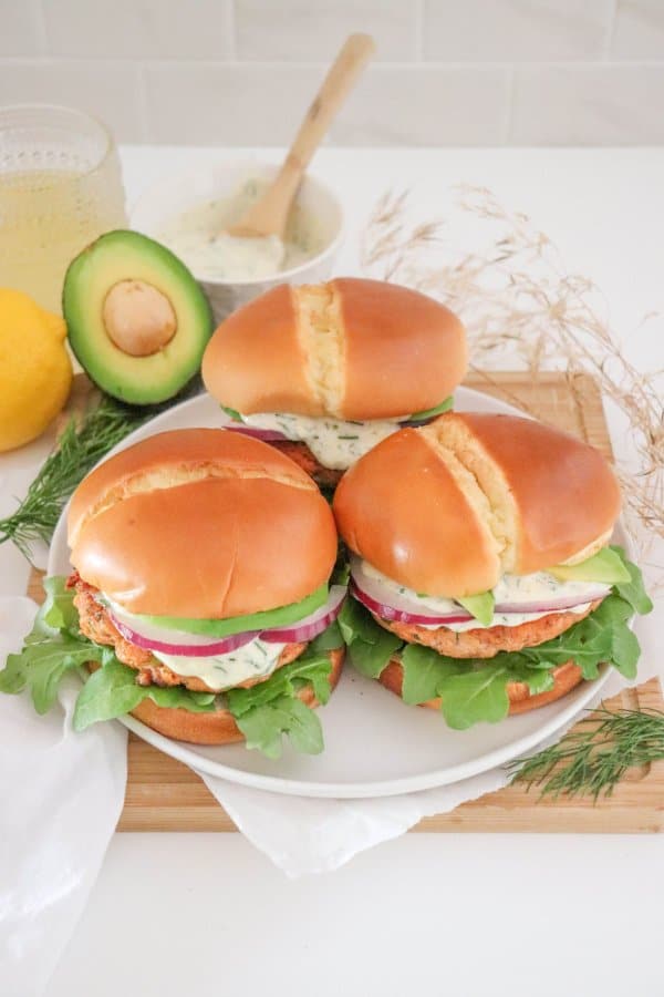Three salmon burgers topped with creamy dill sauce, red onion and avocado on a bed of lettuce in a white bun on a white plate.