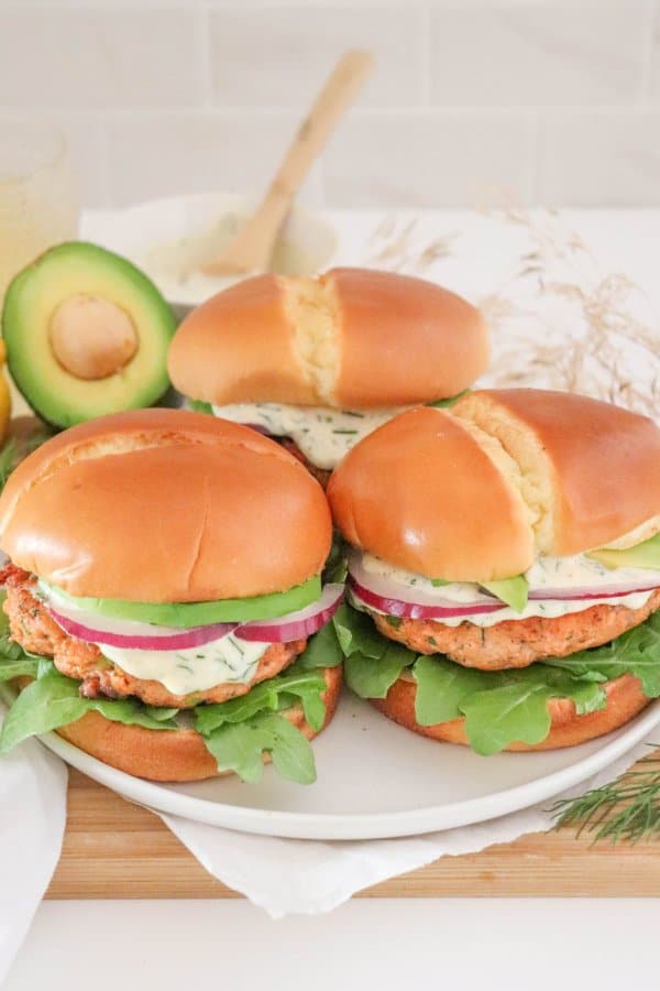 Three salmon burgers topped with creamy dill sauce, red onion and avocado on a bed of lettuce in a white bun on a white plate.