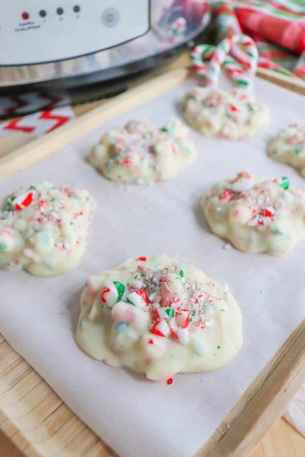 Close up of white chocolate clusters with candy cane sprinkles on a wood tray lined with parchment paper with a crockpot in the background.