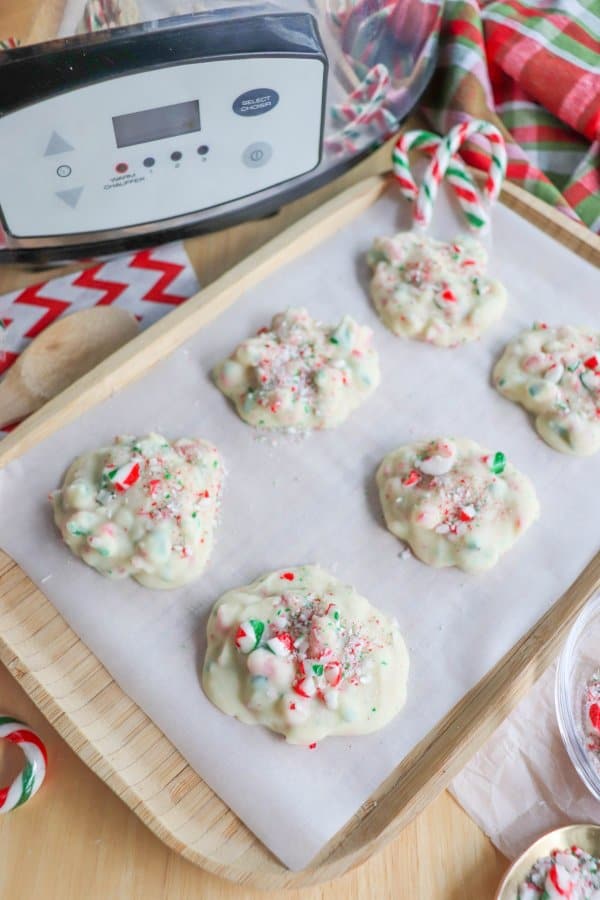 White chocolate clusters with candy cane sprinkles on a wood tray lined with parchment paper with a crockpot in the background.